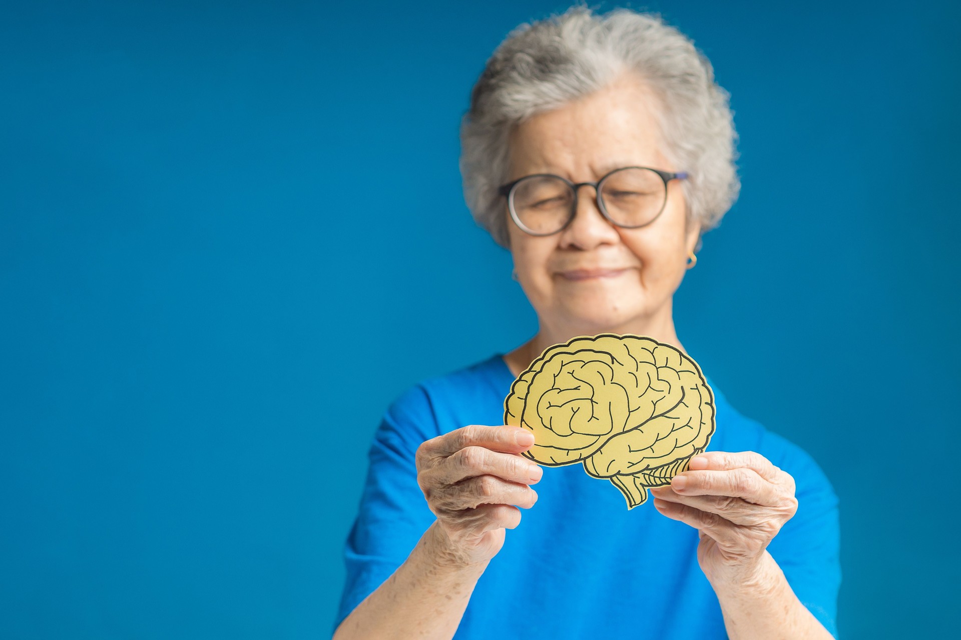 Hands of a senior woman holding a brain shape made from yellow paper against a blue background